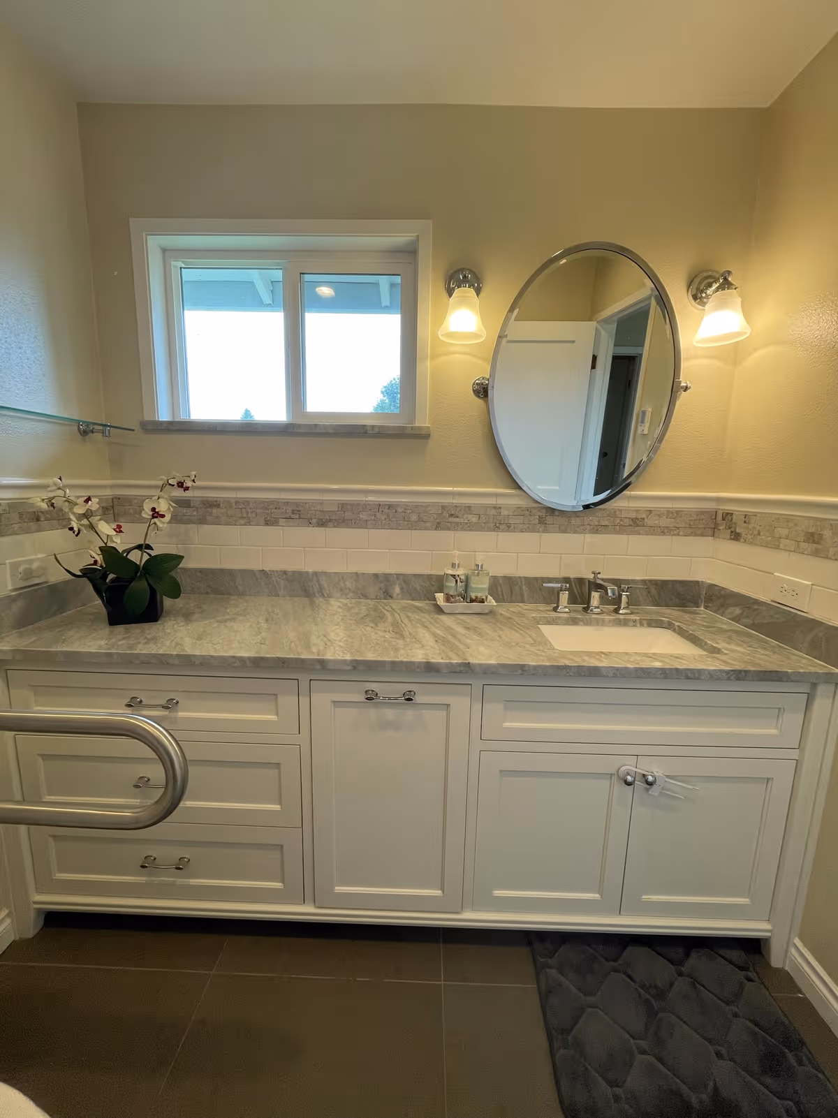Well-lit bathroom vanity with a marble countertop, oval mirror, wall sconces, potted orchid, and a safety grab bar.