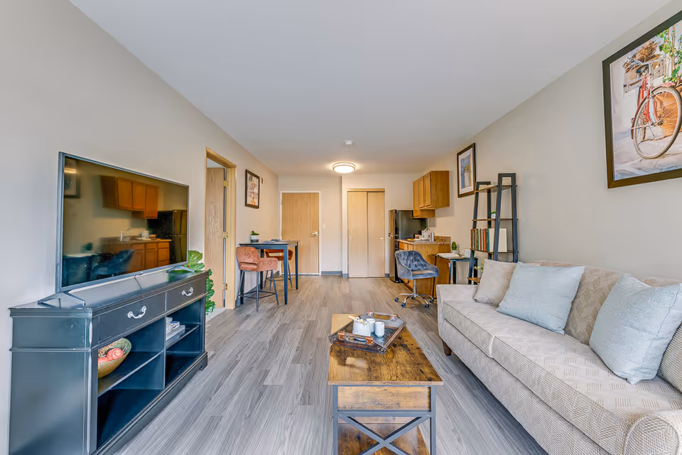 A bright and modern senior living apartment featuring a beige patterned sofa with light blue cushions on the right, a wooden coffee table with a tray and cups in the center, and a black TV stand with a flat-screen TV on the left. The room has light gray wood flooring, neutral-colored walls, and framed artwork. In the background, there is a small dining area with two chairs and a table, a kitchenette with wooden cabinets, a refrigerator, and a desk with a blue chair. Two wooden doors are visible at the far end of the room.
