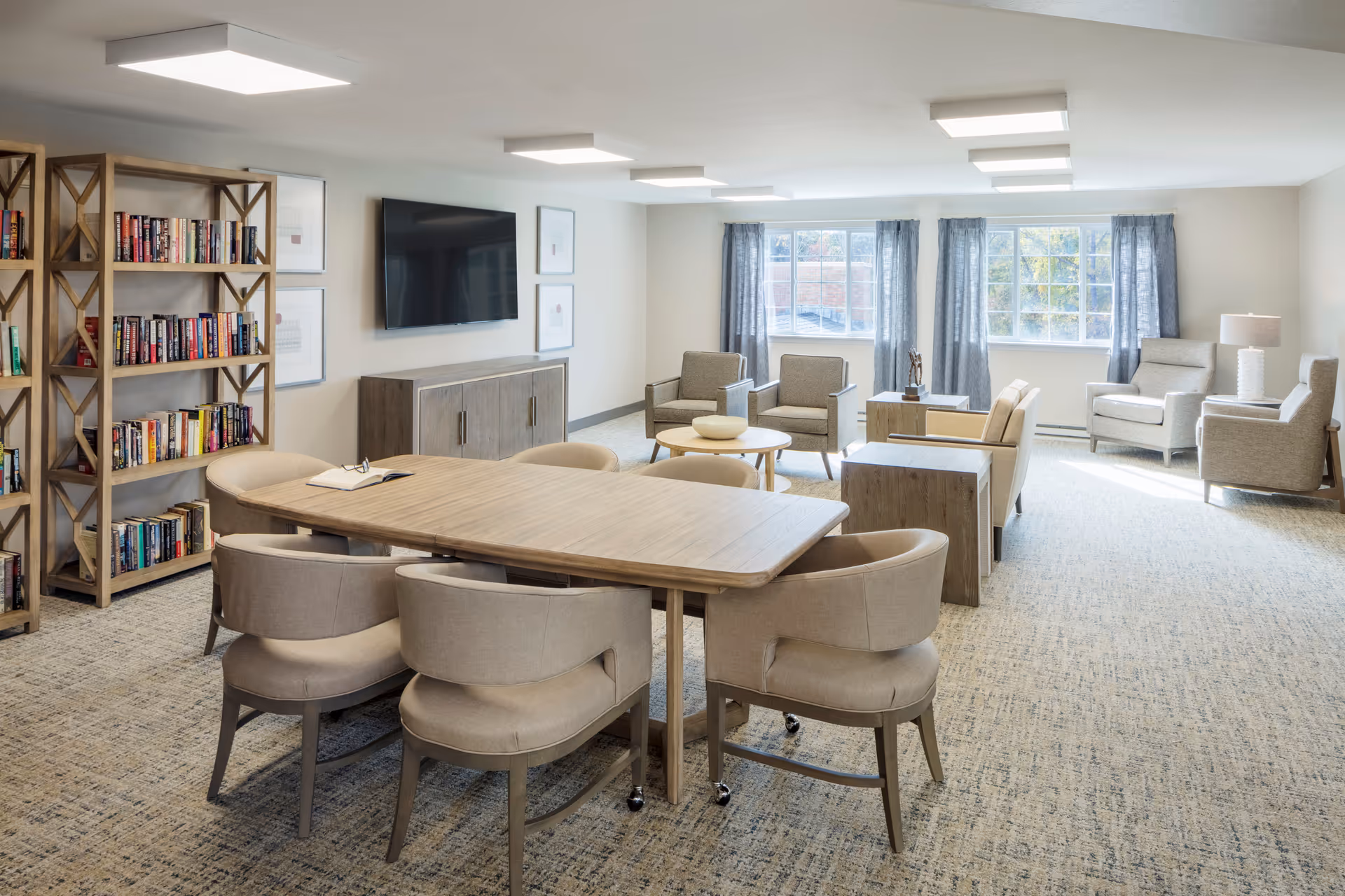A bright and spacious common area in a senior living facility featuring a large wooden table with six beige upholstered chairs, a bookshelf filled with books, a wall-mounted flat-screen TV, and several comfortable armchairs arranged near large windows with gray curtains allowing natural light to fill the room.