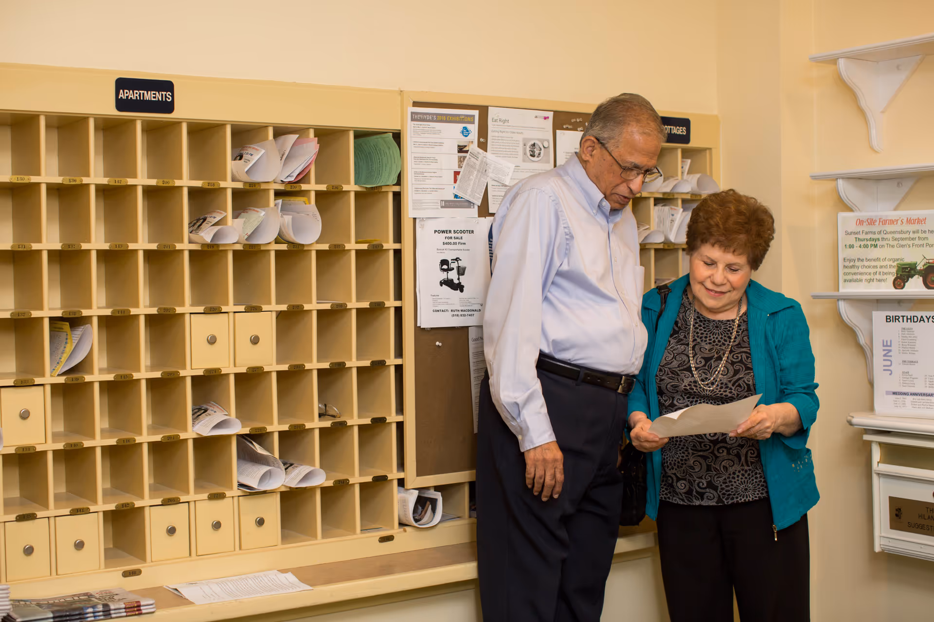 An elderly man and woman standing together in front of a mail sorting area with cubbyholes labeled for apartments and cottages. They are looking at a piece of paper, and there are various notices and flyers pinned on a bulletin board behind them.