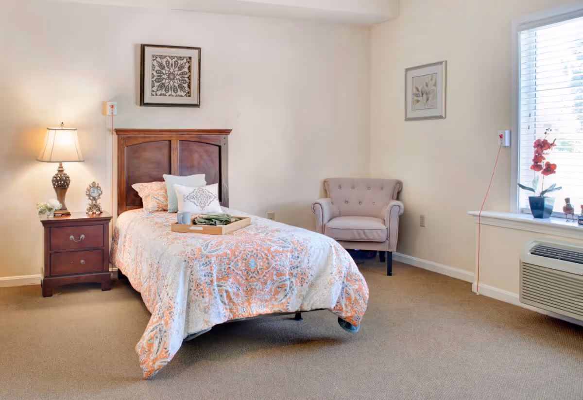 A cozy bedroom with a single bed covered in a patterned orange and white bedspread. There are several pillows on the bed and a wooden tray with a cup and napkin. To the left of the bed is a wooden nightstand with a decorative lamp, a small clock, and a flower arrangement. On the right side of the room, there is a cushioned armchair and a window with blinds, a potted plant, and an air conditioning unit below the window. The walls are light-colored with framed artwork hanging.