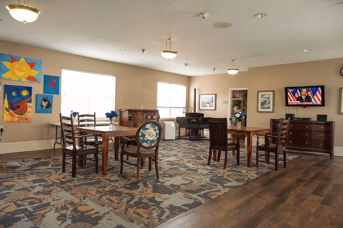 A spacious common area with wooden tables and chairs arranged on a patterned carpet. The walls are decorated with colorful artwork and framed pictures. There is a flat-screen TV mounted on the wall showing a news broadcast. The room has large windows letting in natural light and ceiling lights providing additional illumination.
