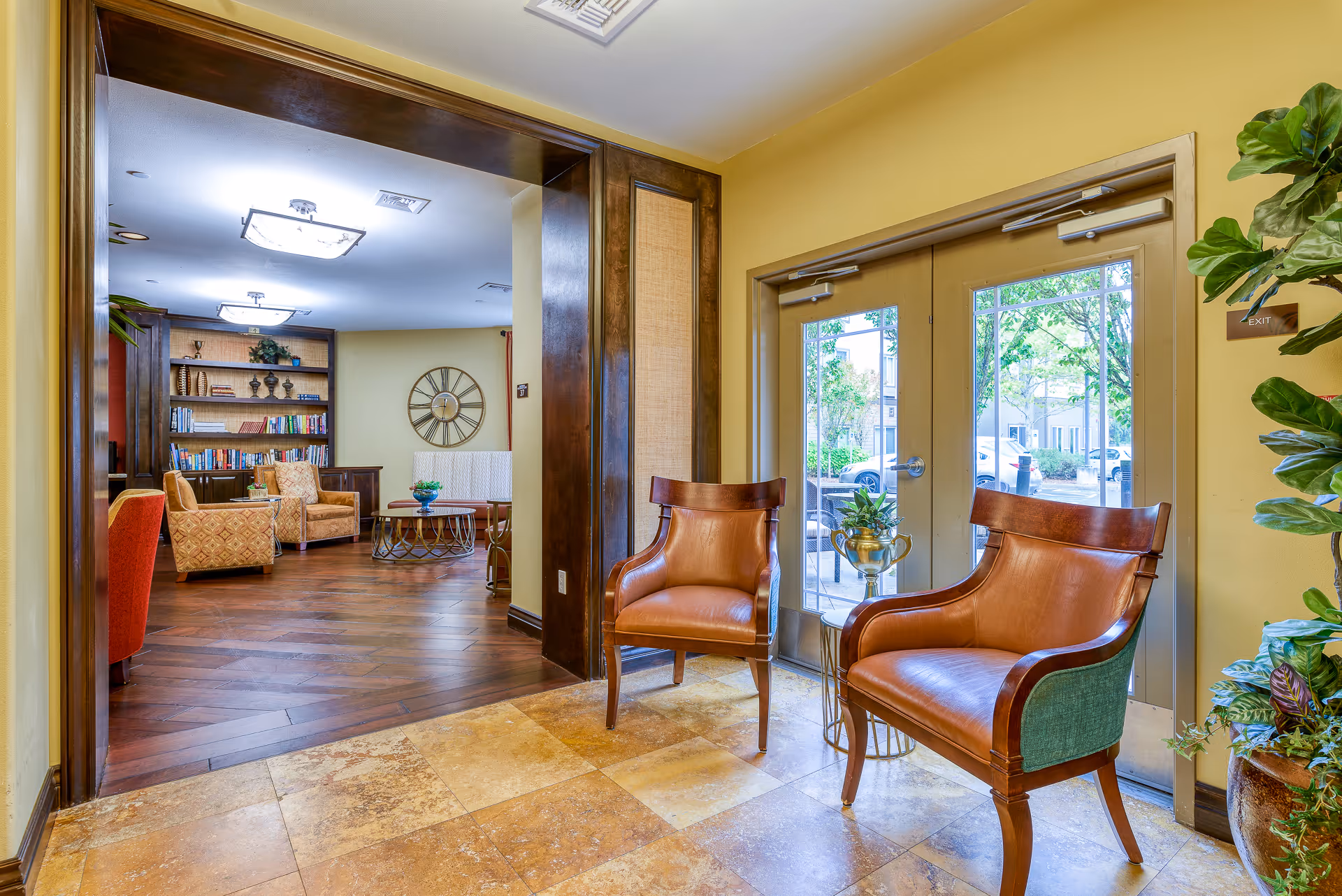 Interior view of a senior living facility lounge area with two brown leather chairs near glass double doors leading outside. The room has yellow walls, tiled flooring near the doors, and wooden flooring in the lounge area. The lounge features armchairs, a coffee table, a large wall clock, and built-in bookshelves with books and decorative items.