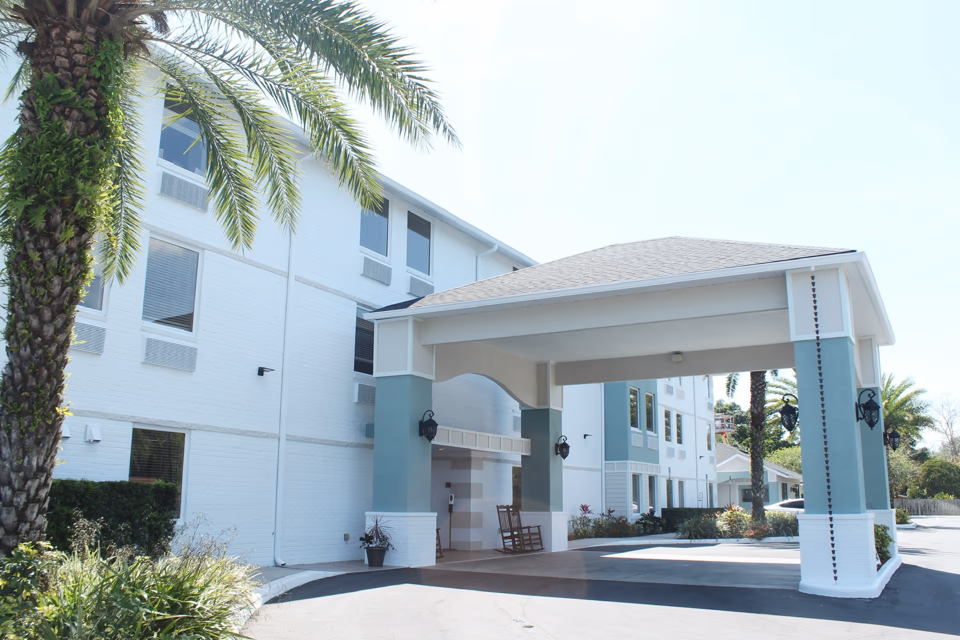 Front entrance of a white three-story senior living building with a covered porte-cochere and palm trees.
