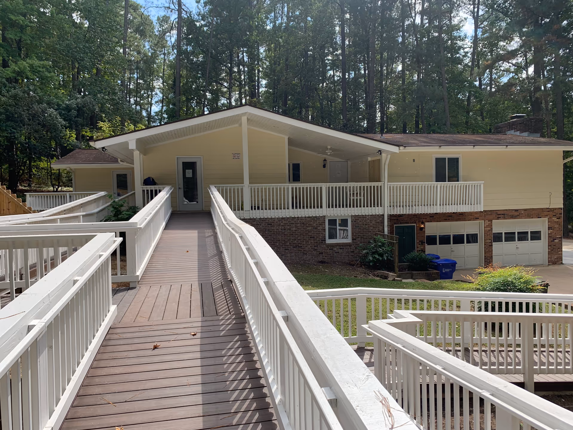 Exterior view of a single-story building with a beige upper facade and brick lower facade surrounded by tall trees. The building has a covered porch area and a wooden ramp with white railings leading up to the entrance. There are two garage doors and a green door visible on the lower level.