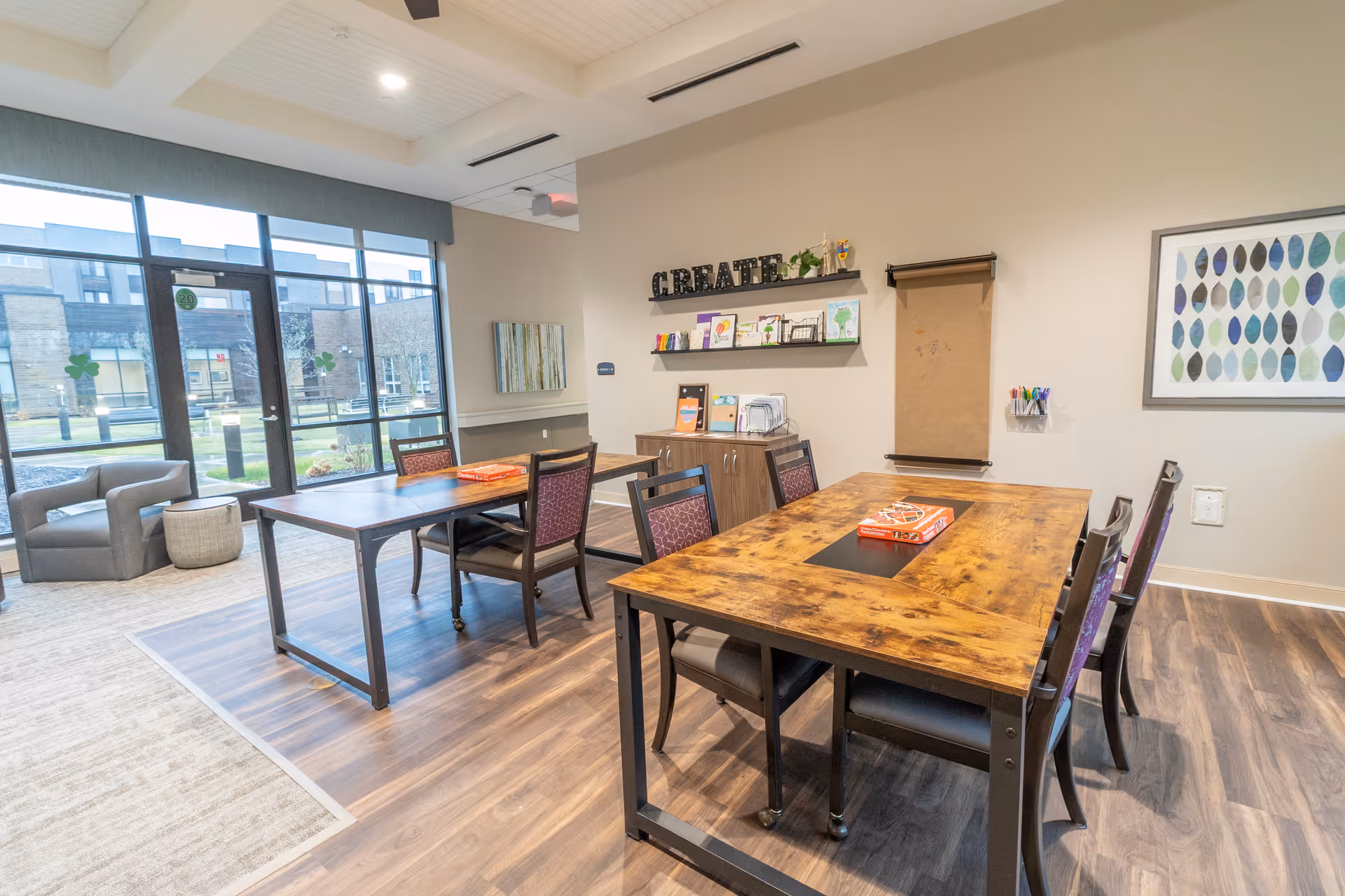 A bright and inviting activity room with wooden tables and chairs arranged for group activities. The room features large windows letting in natural light, a wall-mounted shelf with art supplies and decorations, a roll of brown craft paper for drawing or writing, and a framed abstract leaf artwork on the wall. The floor is a combination of wood and carpet, and there is a comfortable armchair near the windows.