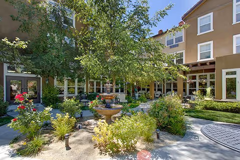 A sunny outdoor courtyard area at The Seasons of Reno featuring a central water fountain surrounded by green shrubs, flowering plants, and trees. The courtyard is enclosed by a multi-story building with large windows and a patio area with seating.