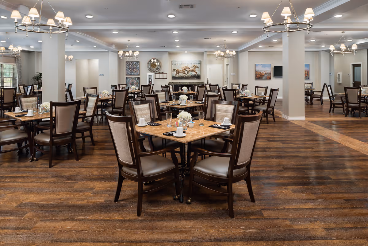 A spacious senior living dining room with multiple wooden tables and chairs arranged neatly. Each table is set with white cups, glasses, napkins, and small floral centerpieces. The room features wooden flooring, soft lighting from chandeliers, and artwork on the walls, creating a warm and inviting atmosphere.