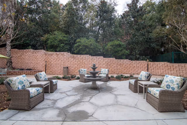 Outdoor patio area with six wicker chairs featuring floral cushions arranged around a central stone fountain. The patio is paved with large stone tiles and is bordered by a low brick wall with trees and greenery in the background.