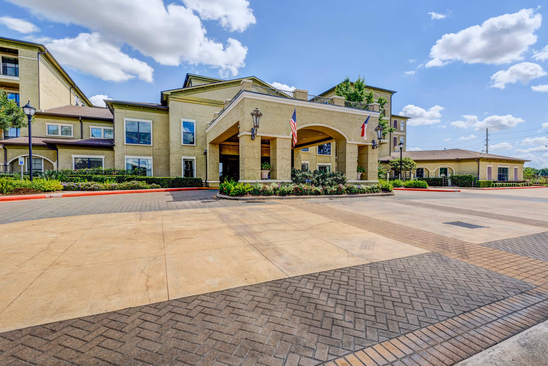 Front entrance of the Conservatory At Champion Forest building with a porte-cochere, flags, landscaping, and a paved driveway under a blue sky.