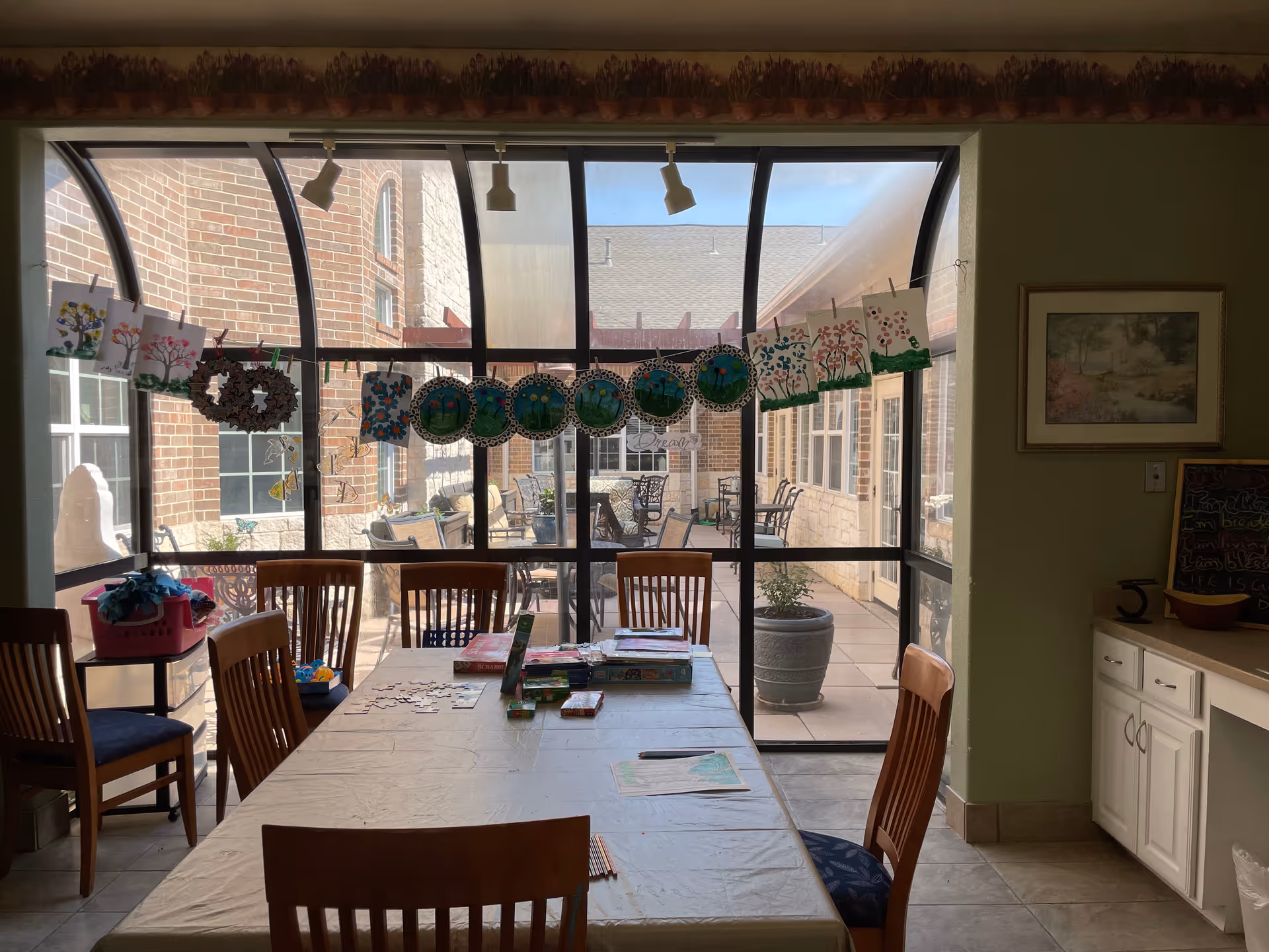 A room with a large table covered with a white tablecloth and several wooden chairs around it. On the table are puzzles, coloring pages, and colored pencils. The room has a large window with a curved glass roof, showing an outdoor patio with tables and chairs. Various colorful artworks and crafts are hanging on a string across the window. There is a cabinet with drawers and a framed picture on the wall to the right.
