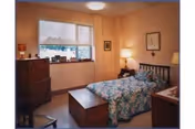 A cozy bedroom with a single bed covered in a floral bedspread, a wooden chest at the foot of the bed, a wooden dresser with framed photos and decorations, a window with a cityscape view, and warm lighting from a table lamp and ceiling light.