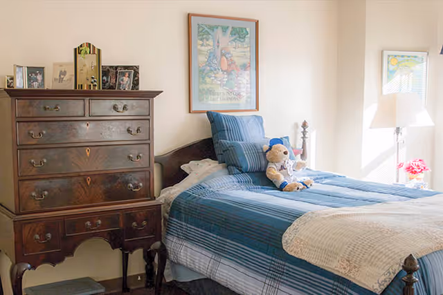 A cozy bedroom with a wooden bed featuring blue striped bedding and a teddy bear sitting on the bed. Next to the bed is a wooden chest of drawers with framed photos on top. The room is softly lit by natural light coming through a window with a white lamp and a small vase of red flowers on the windowsill. Two framed pictures hang on the walls.