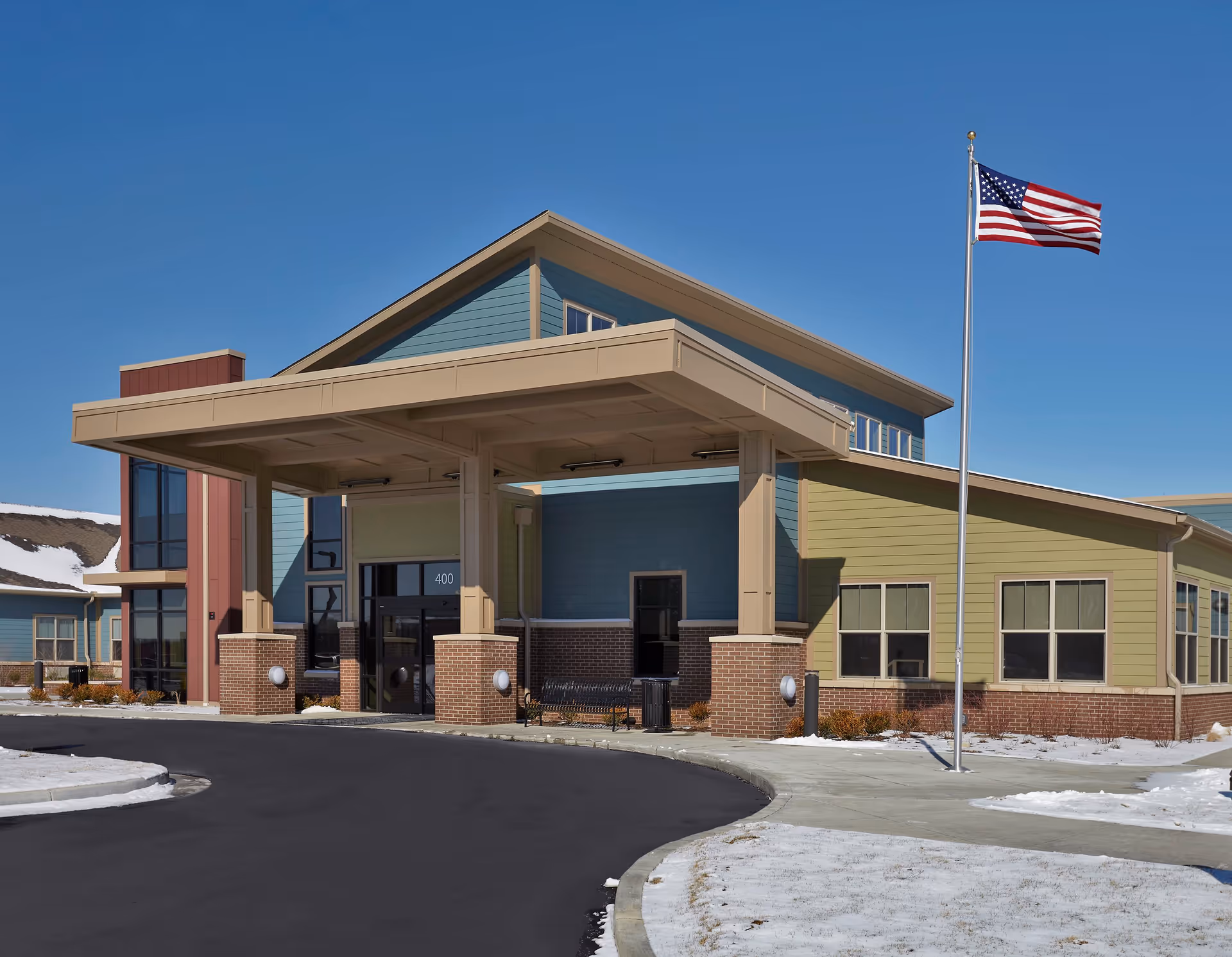 Exterior view of The Springs of Richmond senior living facility building with a covered entrance, brick and siding facade, an American flag on a flagpole, and a clear blue sky. Snow is visible on the ground around the paved driveway.