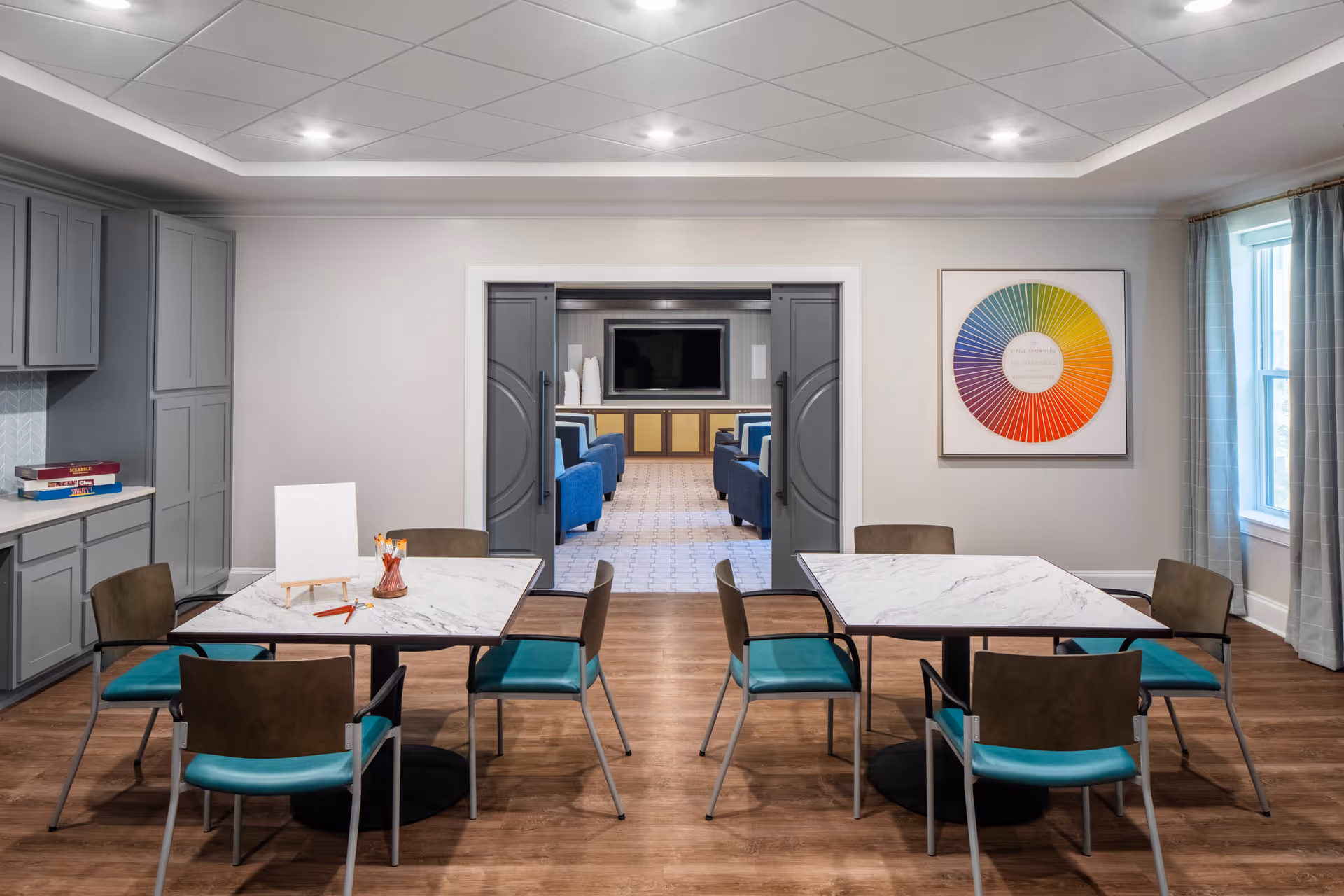 A bright, modern room with two square marble tables, each surrounded by four chairs with teal seats and brown backs. The room has wood flooring, gray cabinetry on the left side, and a colorful circular artwork on the right wall. Through double gray doors, a lounge area with blue chairs and a large TV is visible.