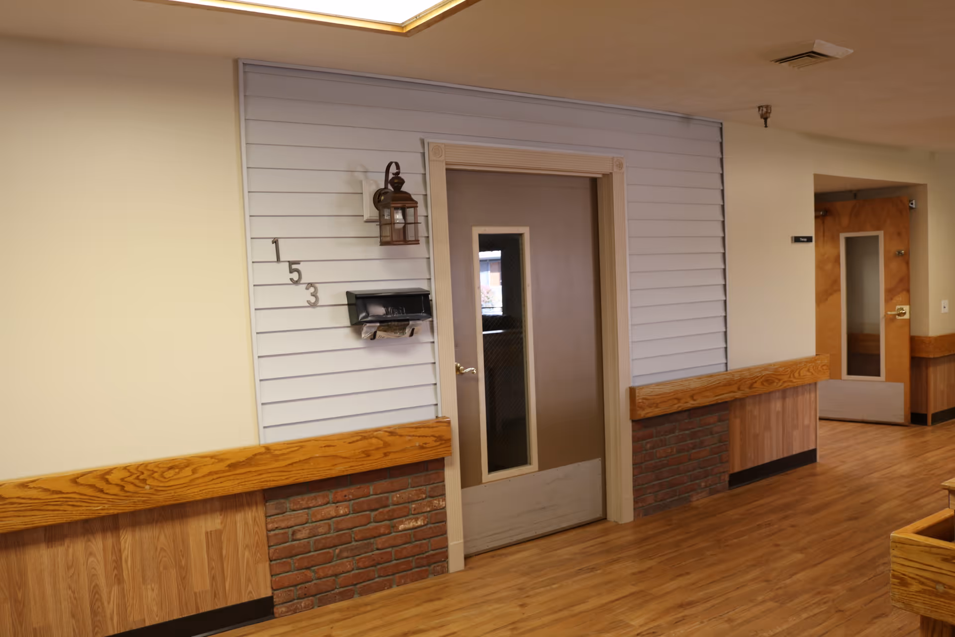 Indoor hallway with wooden flooring and walls partially covered with wood paneling and brick. There are two doors visible, one with the number 153 and a mailbox attached, and a lantern-style light fixture above it. The other door is further down the hallway with a window panel. The ceiling has a recessed light panel.