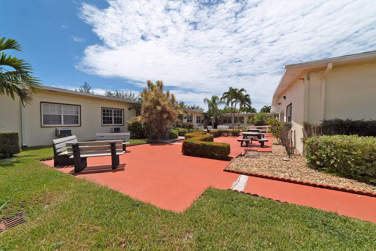 Courtyard with red paved walkways, benches and picnic tables between single-story buildings and landscaped greenery under a partly cloudy sky.
