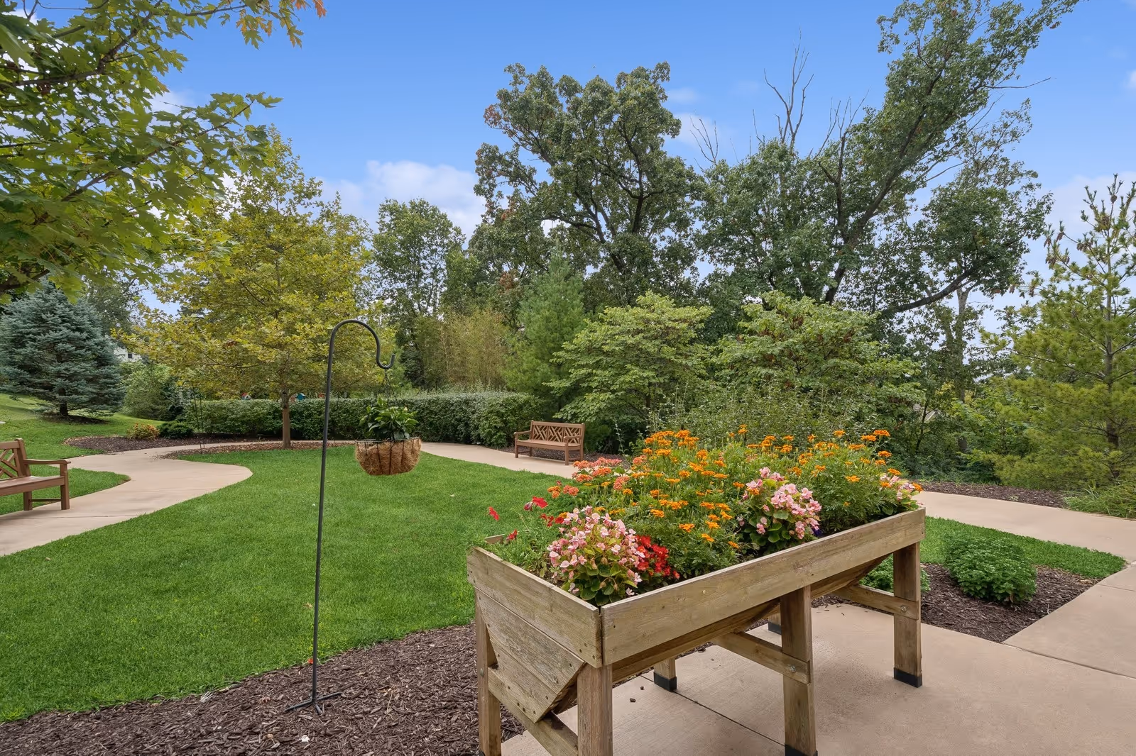 A well-maintained outdoor garden area with a raised wooden planter filled with colorful flowers, green grass, paved walking paths, wooden benches, and various trees and shrubs under a clear blue sky.