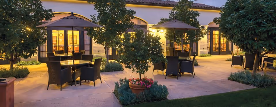Outdoor patio area at dusk with multiple round tables and chairs under large umbrellas, surrounded by small trees and plants, with a building featuring large windows and doors illuminated from inside in the background.