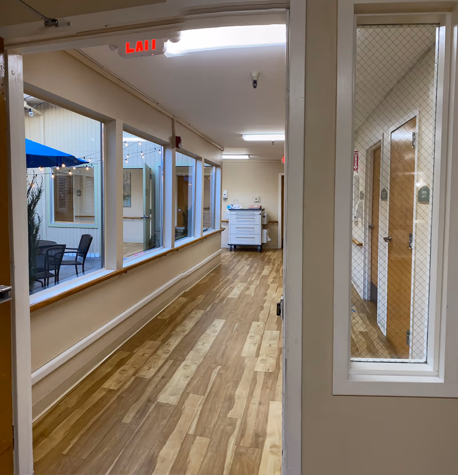 Well-lit hallway in a senior care facility with wood-look floors, windows overlooking a small courtyard with patio furniture, and a medical cart at the far end.