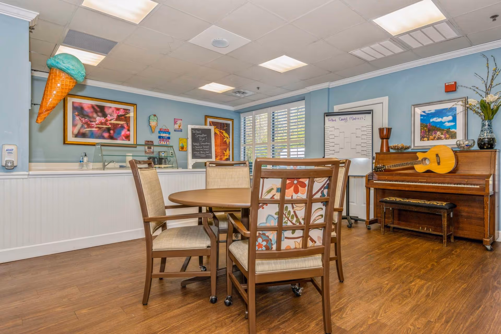 A cozy common area with a round wooden table surrounded by four chairs, one with a floral patterned back. The room has light blue walls and wood flooring. On the right side, there is an upright piano with a guitar resting on top and a vase with flowers. A whiteboard with a bracket titled 'Talbot Candy Madness!' is visible near the piano. On the left side, there is a counter with a glass sneeze guard and a large decorative ice cream cone hanging on the wall above it. Various colorful signs and framed pictures decorate the walls.
