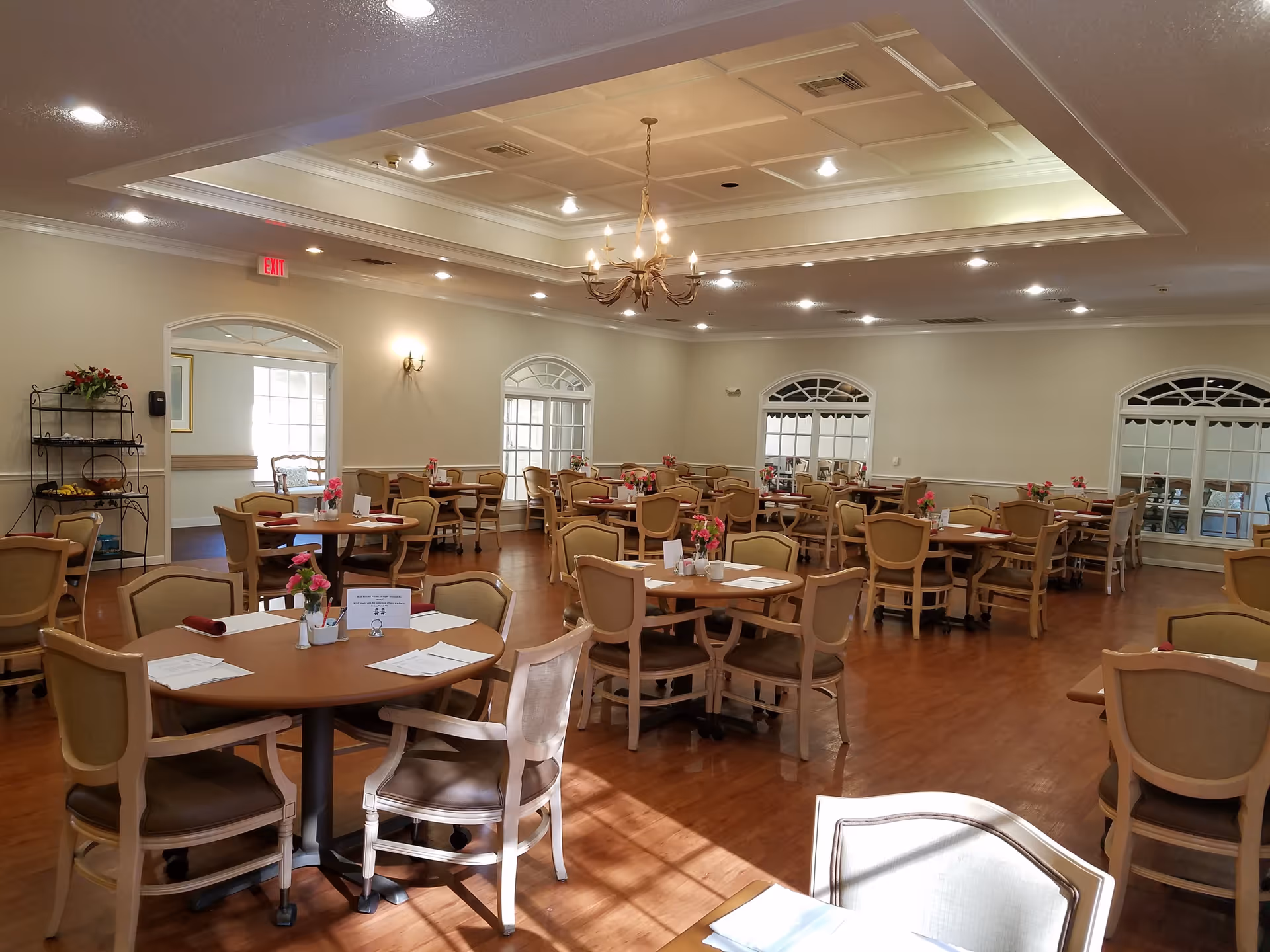 A spacious dining room with multiple round tables and beige cushioned chairs arranged neatly. Each table has a small flower vase with pink flowers and place settings with napkins and menus. The room features a coffered ceiling with recessed lighting and a chandelier in the center. Large windows with decorative arches allow natural light to enter the room. The floor is wooden, and there is a metal shelving unit with fruit and other items near an arched doorway.