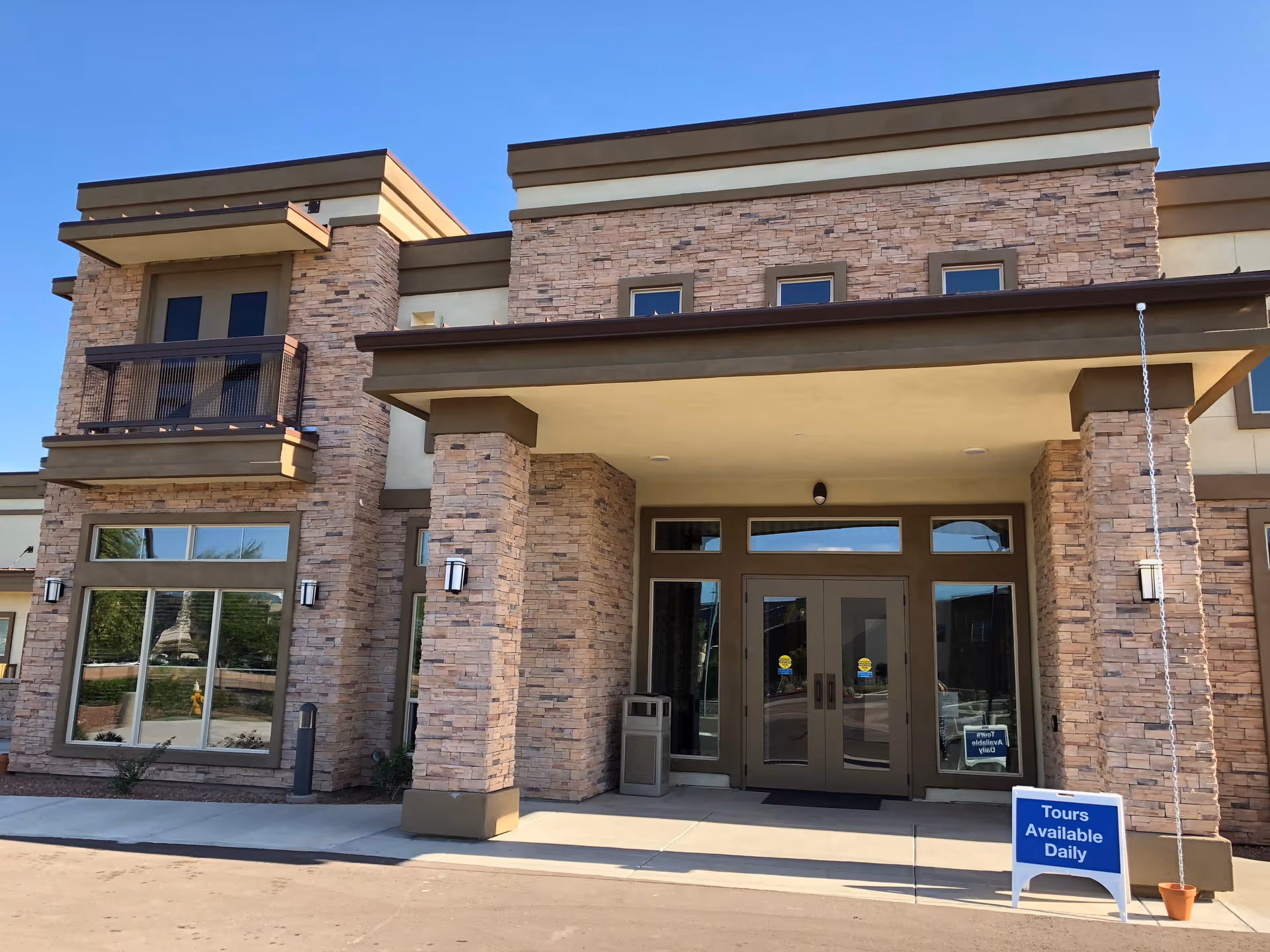 Front exterior view of a modern building with stone and beige stucco facade, large windows, double glass doors, and a covered entrance supported by two stone pillars. A blue sign near the entrance reads 'Tours Available Daily.'
