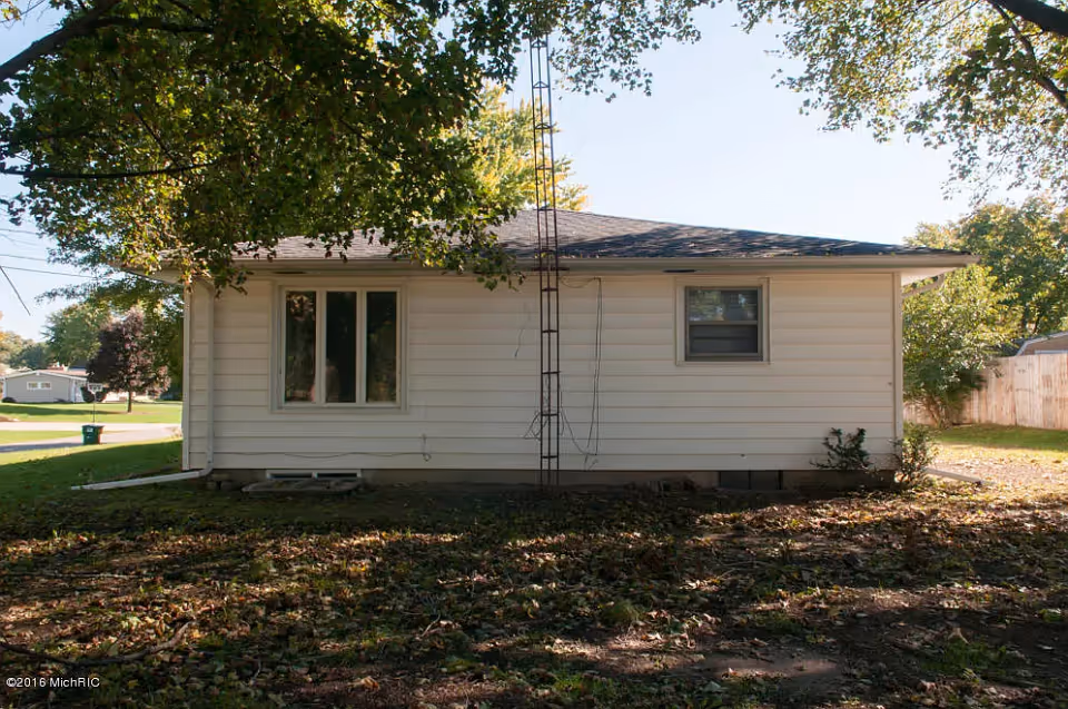 Single-story white-sided house exterior with two windows, a metal antenna and trees casting shade over the yard.
