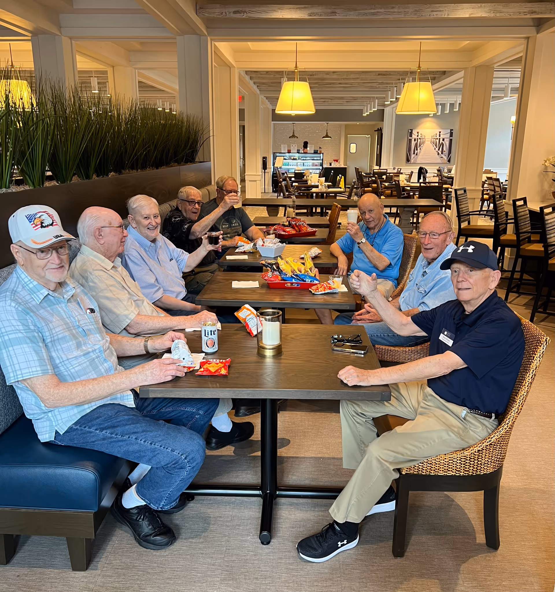 A group of eight elderly men sitting around two tables in a dining area, enjoying snacks and drinks. The room is well-lit with hanging lamps, and there are more tables and chairs in the background along with a display counter. The men appear to be socializing and having a good time.