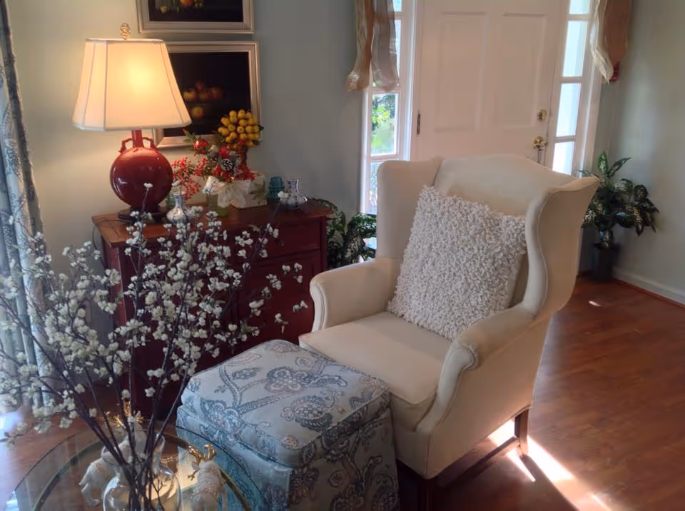 A cozy living room corner featuring a cream-colored wingback armchair with a textured white pillow, a patterned ottoman in front of it, a wooden side table with a red lamp and decorative fruit arrangements, and a glass coffee table with a vase of white flowers. The room has wooden flooring, light-colored walls, and a door with windows on either side letting in natural light.