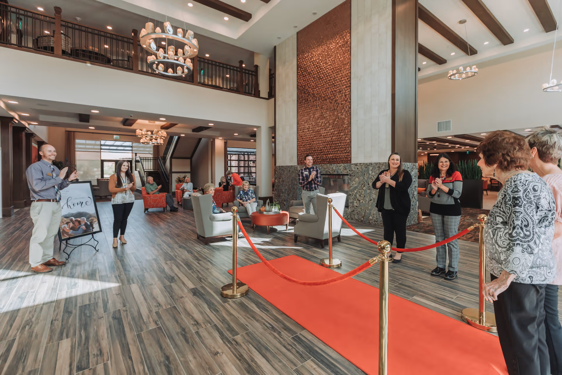Spacious, well-lit senior living lobby with a red carpet and gold stanchions, seating areas, chandeliers, and people gathered and clapping.