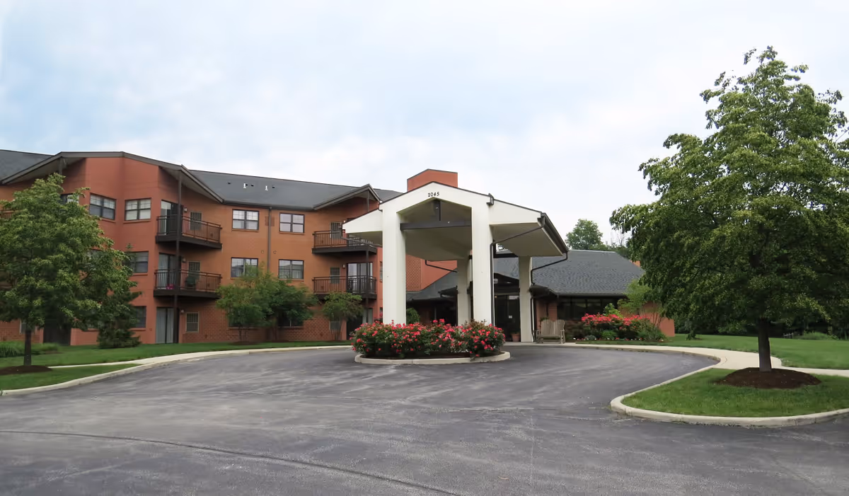 Front entrance of a brick senior living building with a covered porte-cochère, circular driveway, balconies and landscaping.