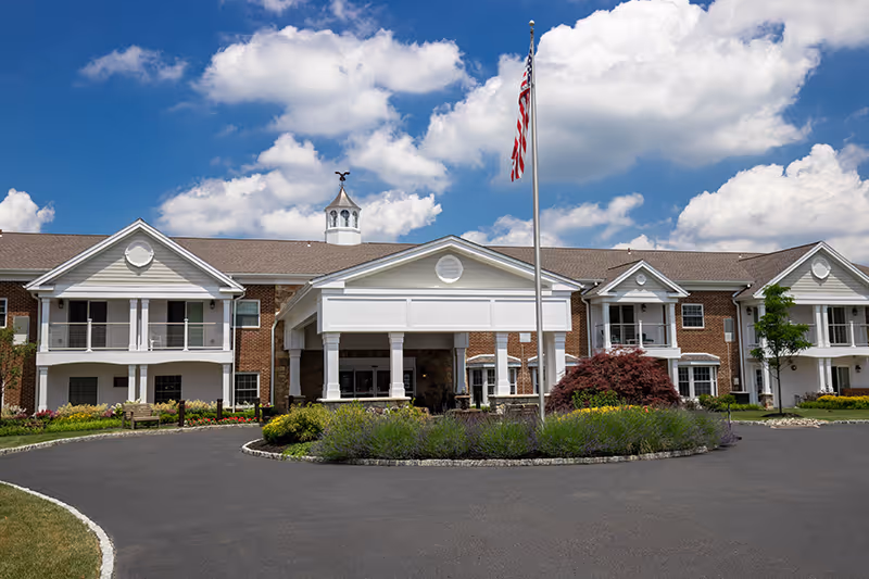 Front entrance of a two-story brick senior living building with a covered porte-cochere, American flag, and landscaped circular driveway under a blue sky.