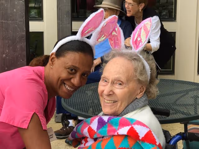 A smiling caregiver and an elderly woman, both wearing pink bunny ear headbands, sitting outdoors at a round metal table. The elderly woman is wrapped in a colorful blanket and seated in a wheelchair. In the background, two other elderly people are seated and engaged in conversation.