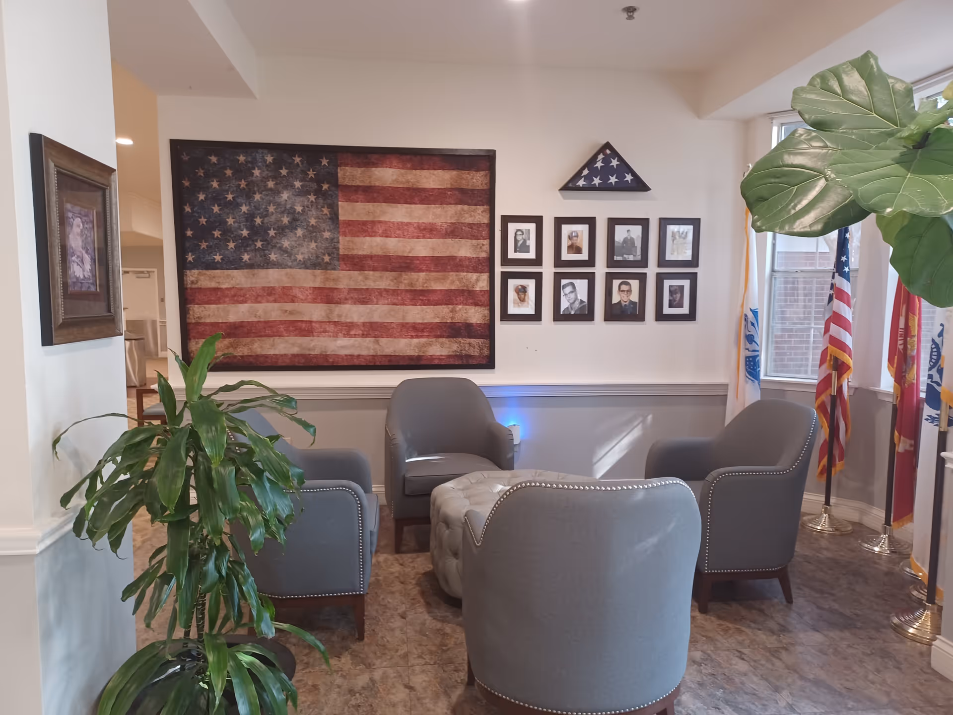 A cozy seating area with four gray armchairs arranged around a tufted ottoman. On the wall behind the chairs is a large vintage American flag, a folded flag in a triangular display case, and eight framed black and white photographs. To the right, several flags stand near windows letting in natural light. A large green plant is visible on the left side of the image.