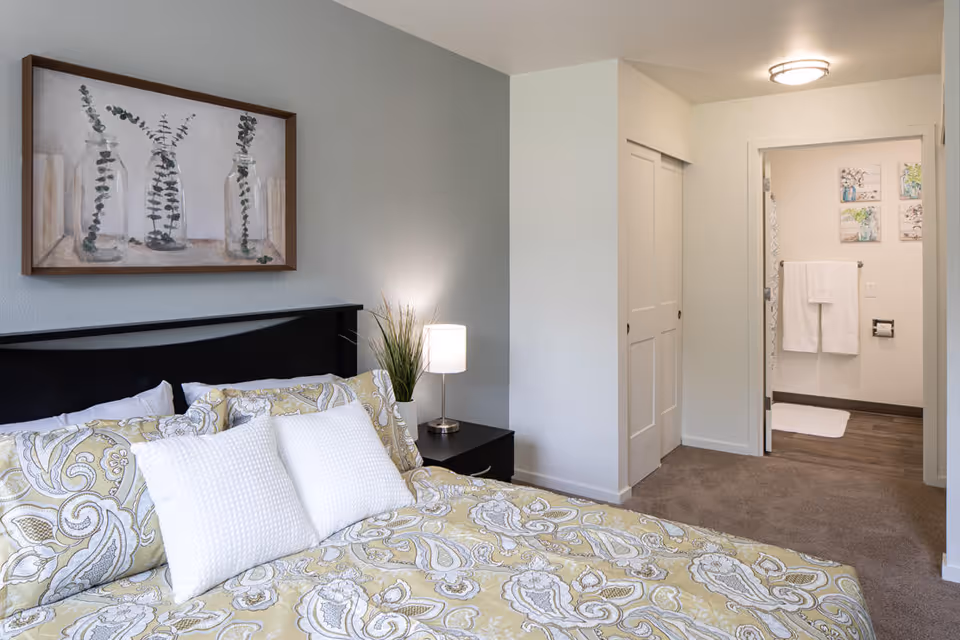 A bedroom with a bed featuring a patterned yellow and white bedspread and two white pillows. There is a black headboard and a nightstand with a lamp and a small plant. On the wall above the bed is a framed artwork of three glass bottles with green foliage. The room has light-colored walls and carpeted floor. In the background, there is an open doorway leading to a bathroom with white towels hanging and decorative wall art.