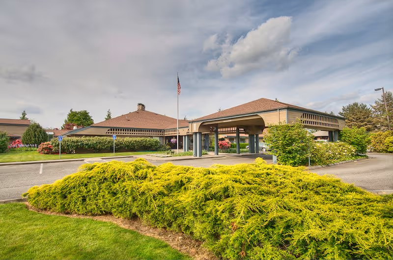 Exterior view of a single-story building with a covered entrance and an American flag on a flagpole. The building is surrounded by well-maintained landscaping including green bushes, trees, and a lawn under a partly cloudy sky.
