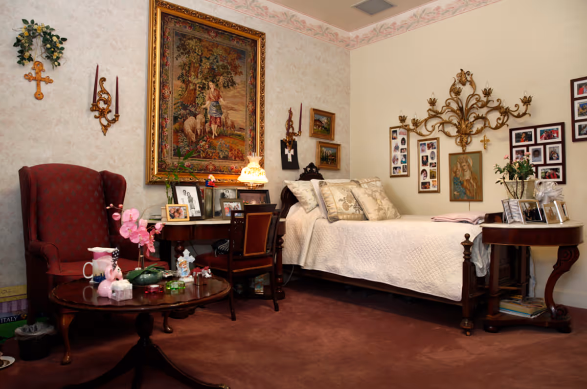 A cozy bedroom with a single bed covered in a white quilt and decorative pillows. The room features a red upholstered armchair, a wooden side table with framed photos and a vase of flowers, and a wooden desk with a chair and a lamp. The walls are decorated with framed pictures, a large tapestry depicting a pastoral scene, and ornate candle holders. The carpet is a deep reddish-brown color.