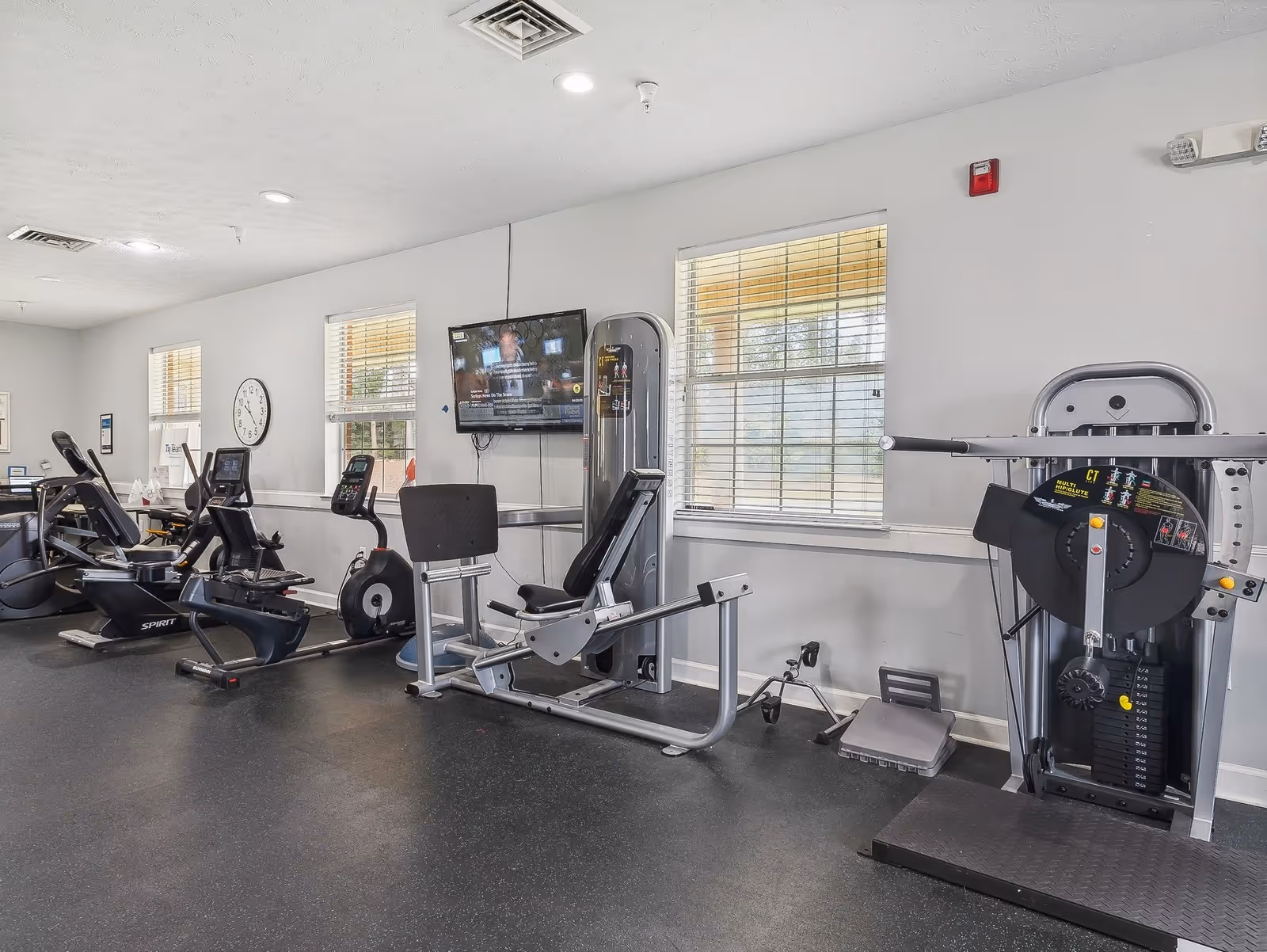 Interior view of a senior living facility's fitness room with exercise equipment including stationary bikes, a leg press machine, and a weight machine. There are windows with blinds, a wall-mounted TV, and a clock on the wall.