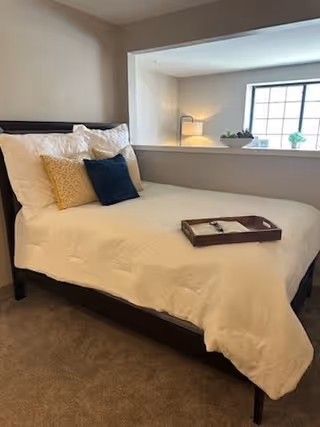 A neatly made bed with white bedding and three decorative pillows in yellow, white, and dark blue. A wooden tray with a notepad and pen rests on the bed. The room has beige walls and carpet, and a large window with a windowsill holding a small plant and decorative items.