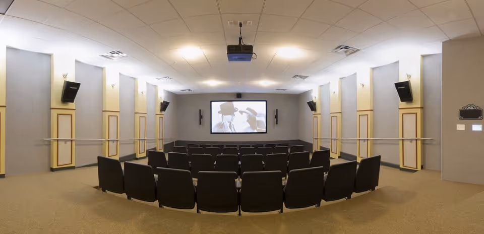 A small theater room with rows of black chairs facing a large screen showing a black and white movie scene with two people wearing hats. The room has beige walls with decorative panels, wall-mounted speakers, and a ceiling-mounted projector.