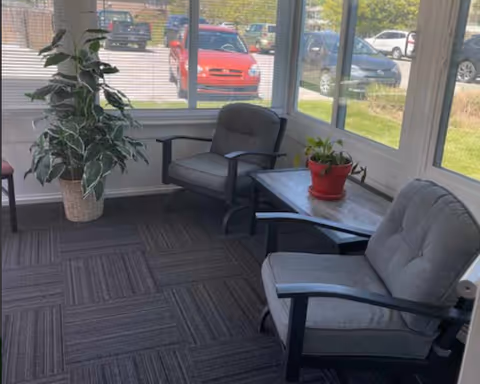 A small sunroom or enclosed porch area with two cushioned armchairs and a small table between them holding a red potted plant. There is a large leafy plant in a woven basket in the corner. Large windows show a parking lot with several cars outside.