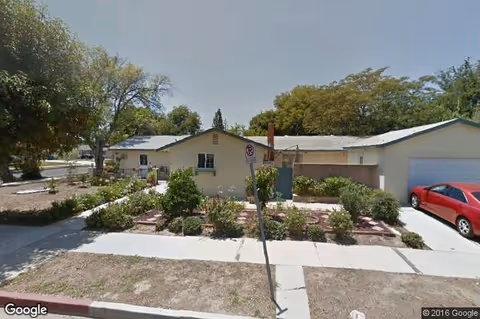 Single-story light-colored building with a landscaped front yard, sidewalk, and a red car parked in the driveway.