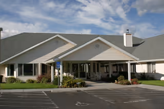 Front exterior view of a single-story senior living facility building with a covered entrance, wheelchair accessible parking space, and landscaped greenery under a partly cloudy sky.