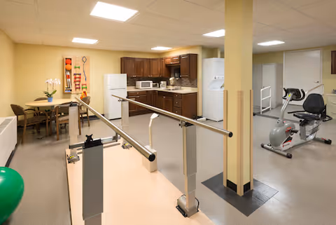 A multi-purpose room featuring a small kitchen area with dark wooden cabinets, a white refrigerator, microwave, and a washing machine. In the foreground, there are parallel bars for physical therapy or rehabilitation. To the right, there is an exercise bike and a white door. On the left side, there is a table with chairs and some colorful wall decorations. The room has beige walls and ceiling lights.