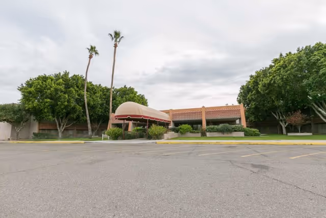 Front exterior of a single-story senior care building with a domed entrance canopy, palm trees, and an empty parking area.