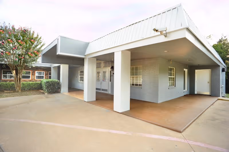 Exterior view of a single-story building with a covered entrance supported by white columns, gray brick walls, and multiple windows. There is a tree and bushes on the left side, and a concrete driveway in front.
