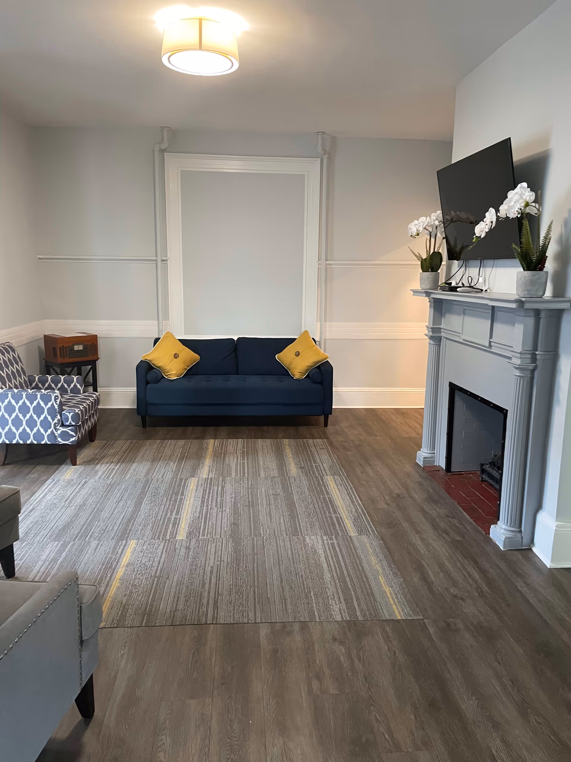 A cozy living room with a blue sofa adorned with two yellow pillows, a patterned armchair to the left, and a gray fireplace on the right with a mounted flat-screen TV above it. Two white orchid plants and a small green plant are placed on the fireplace mantel. The room has light gray walls, a ceiling light fixture, and a carpet with gray and yellow stripes over wooden flooring.