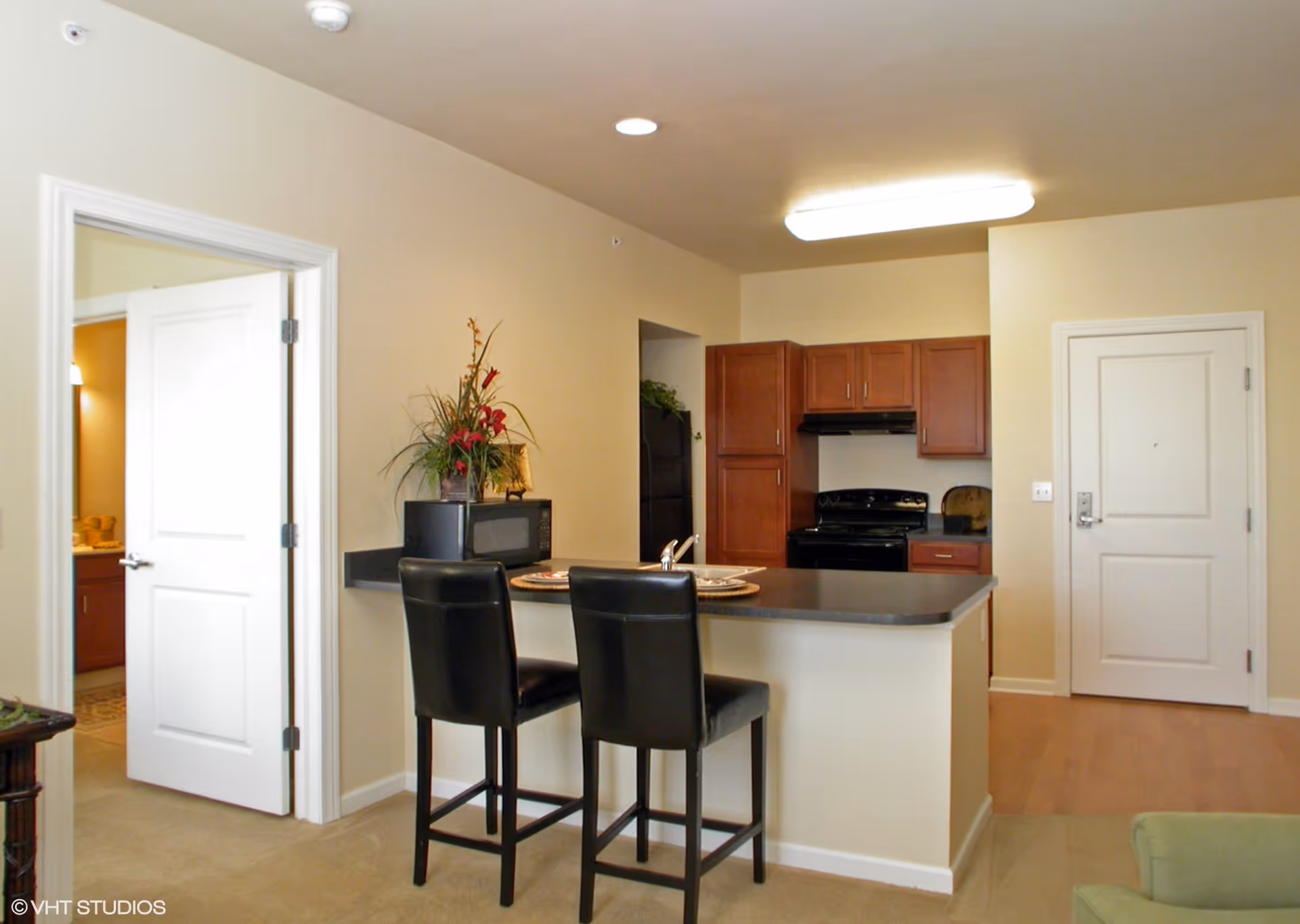 Open-plan kitchen and entry area with a breakfast bar, two black bar stools, wooden cabinets, and a stove.