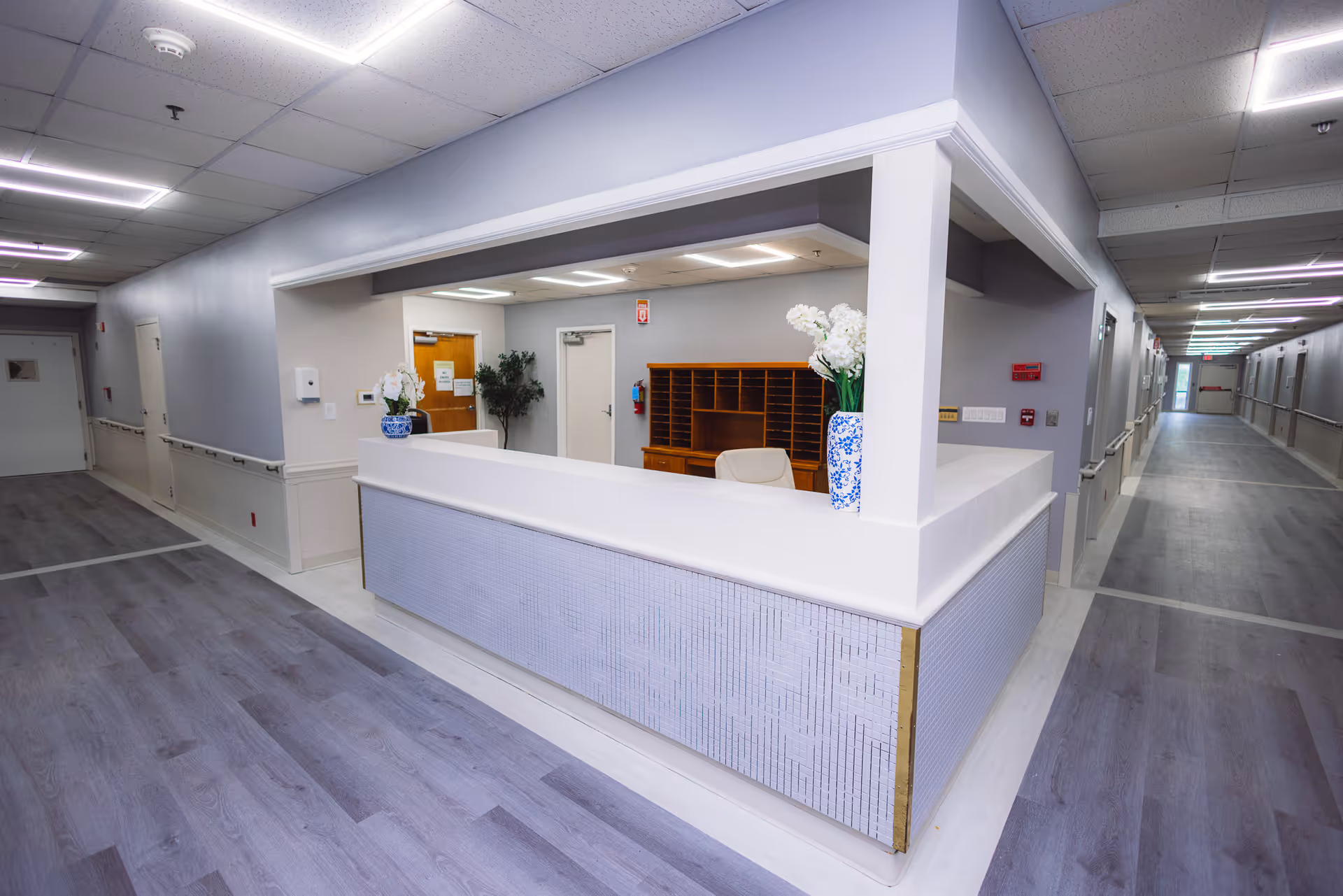 A clean and modern reception desk area in a healthcare facility with light gray walls, wood flooring, and two vases with white flowers on the counter. The desk is situated in a hallway with multiple doors and handrails along the walls.